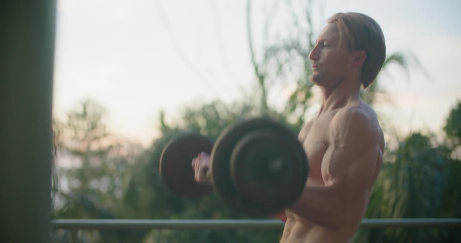Shirtless muscular man lifting weights outside the house at sunset