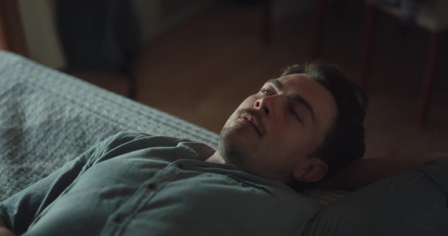 A man in a room laying in bed looking at a smart phone with chairs in the background
