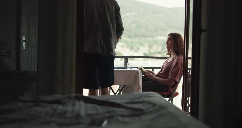 Sad man puts smartphone away and goes up to his girlfriend reading book