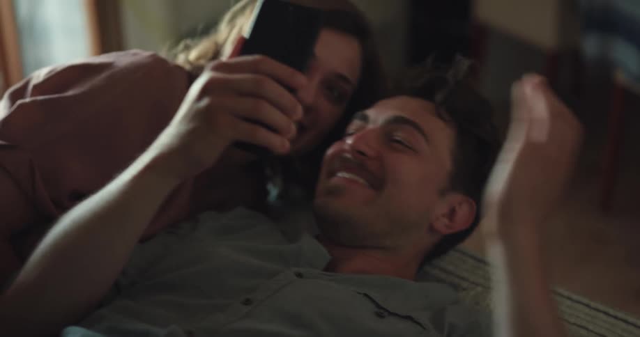 A man and a woman in a room laying in bed and looking at a smart phone