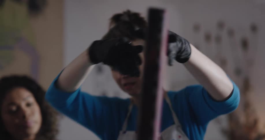 A woman wears gloves while examining a painting as a coworkers watches