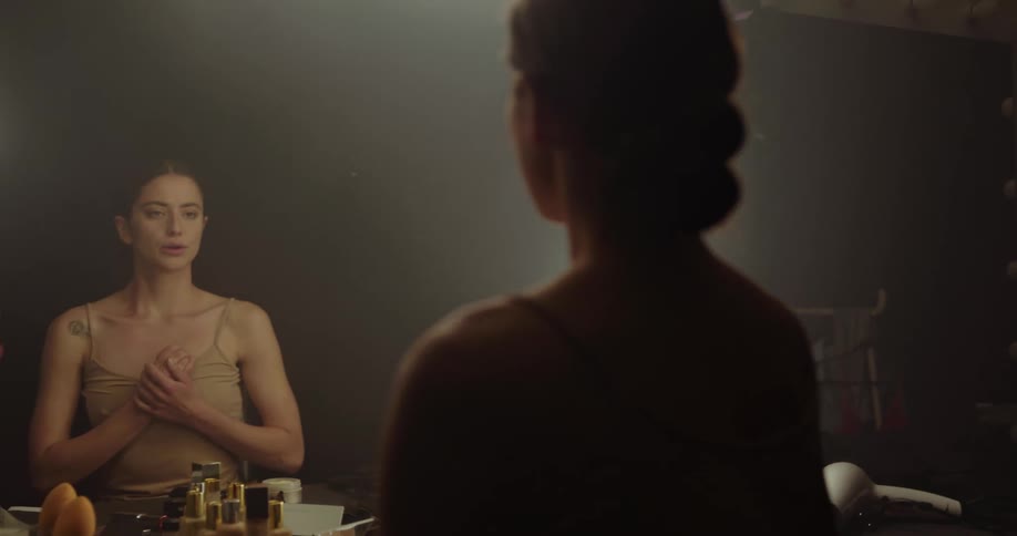 Woman looks at her reflection in a lighted mirror while she sits at a table with several bottles on it