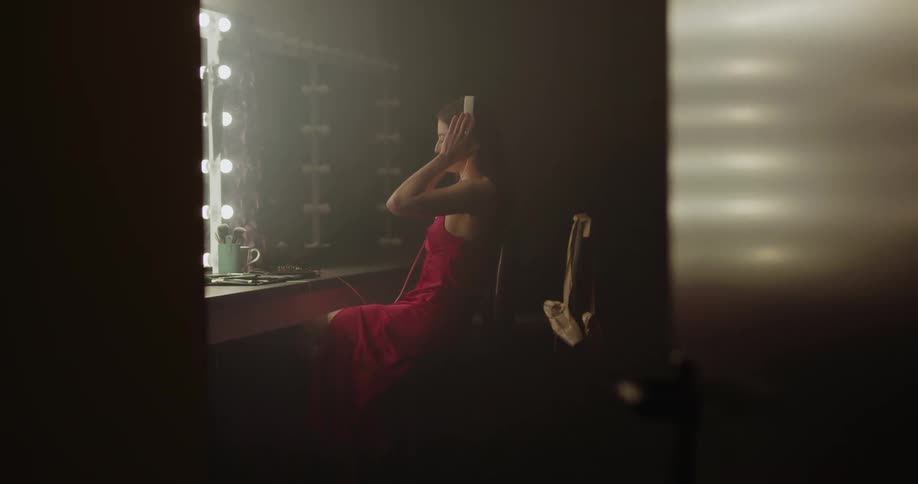 Woman dressed in red dress sits in front of mirror in dressing room listening to music on headphones