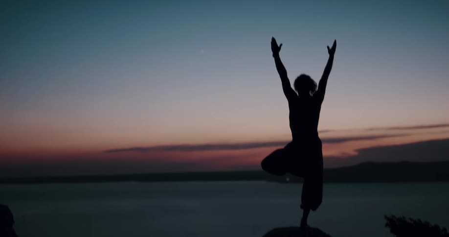 Person silhouetted against red tinted sky stands on one leg in yoga pose overlooking ocean