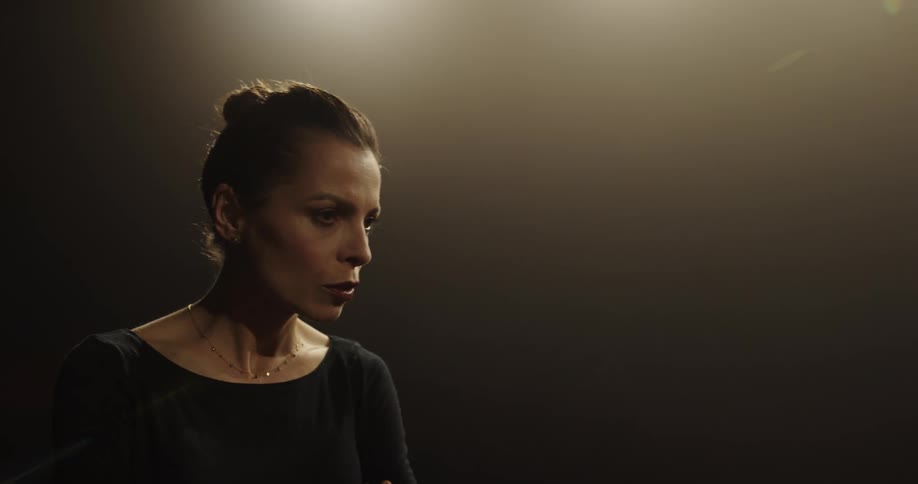 An actress stands under a spotlight and talks and uses her hands as she performs