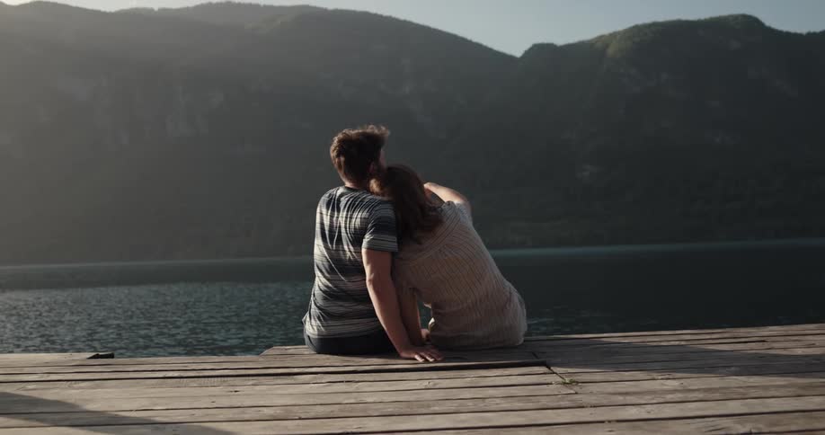 A man and a woman sitting on a dock with a river land the sky and clouds in the background
