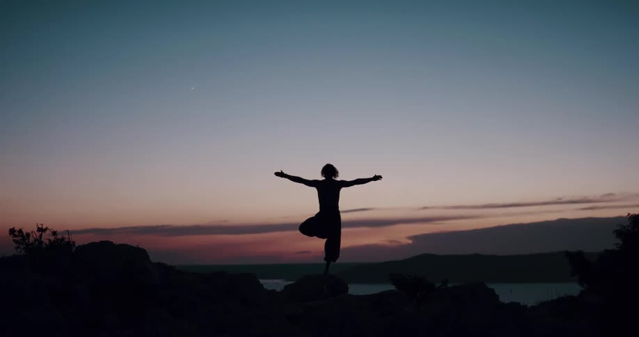 Woman does a tree pose while practicing yoga on a rock near the lake