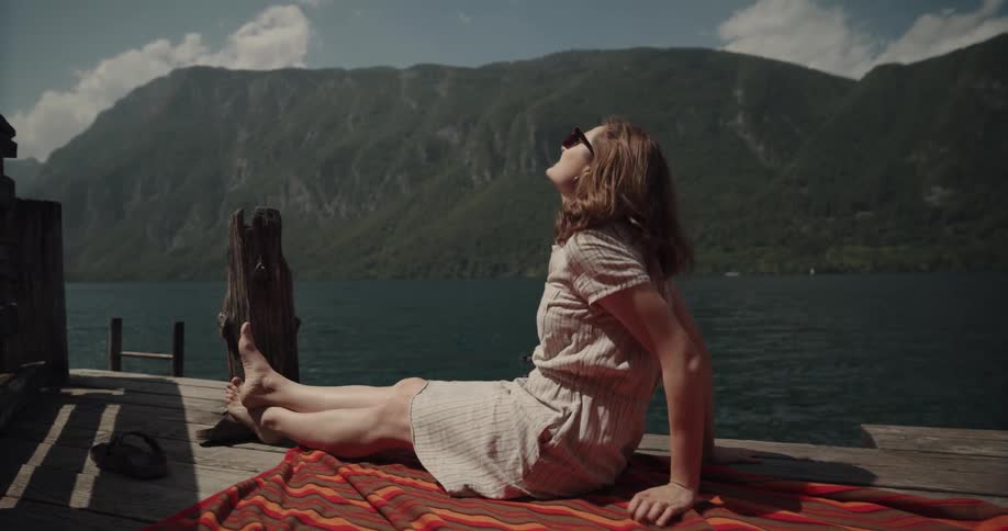 A woman is lounging on a deck on a lake and pushing her hair out of her face as the wind blows