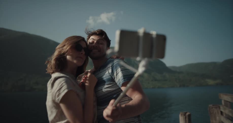 Man holds a selfie stick with a cell phone attached and takes selfies of himself and his girlfriend next to a lake