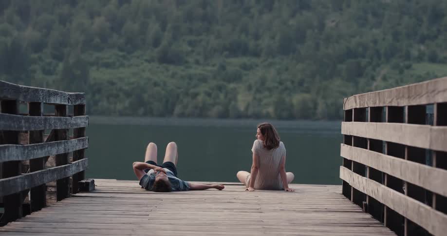 A teenage boy lies on a dock while talking to a teenage girl that sits beside him