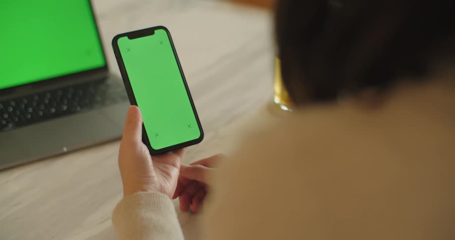 A woman sits at home and holds her smartphone with a solid green screen while a laptop with a green screen sits on a table in front of her