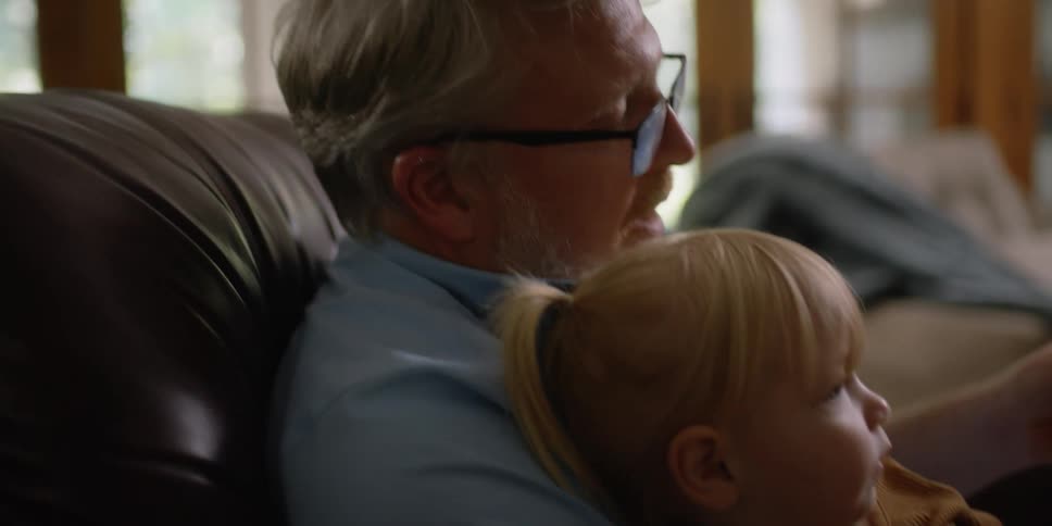 Little child is lying on the lap of her grandfather while he reads her a book