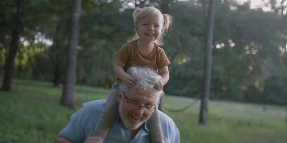 A toddler laughs while swaying back and forth on her grandfathers shoulders in the park