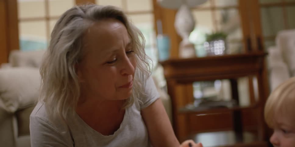 Woman with grey hair sits on the rug with little blonde girl playing with toys
