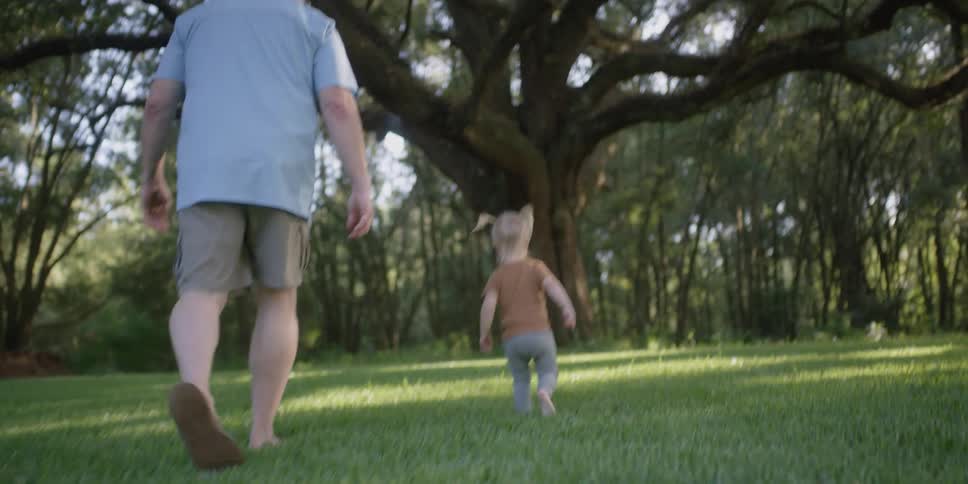 A little girl holds her grandfathers hand as they walk and then she lets go and runs towards a tree as a dog joins them