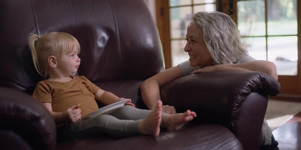 An older woman squats next to a leather chair that a little girl is sitting in with a plate and a cookie