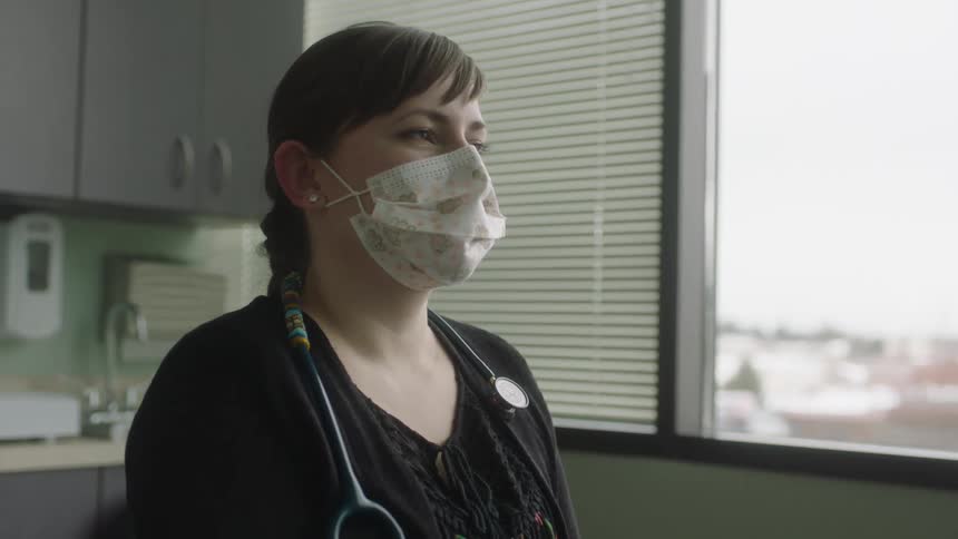 A female doctor wearing a mask is standing in a room in front of a window