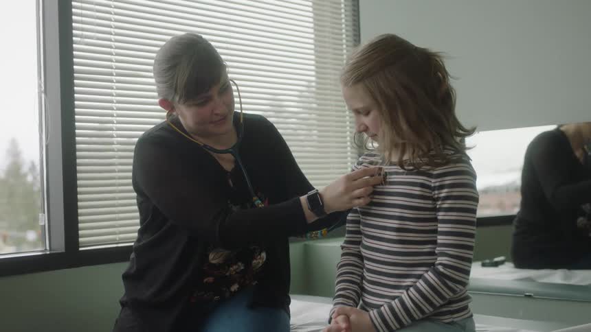 A patient sits on an exam table while a doctor puts a stethoscope on and checks her heartbeat