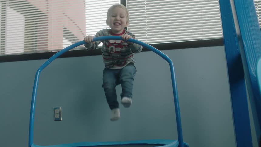 A young boy playfully jumps on a miniature trampoline in a house during the day