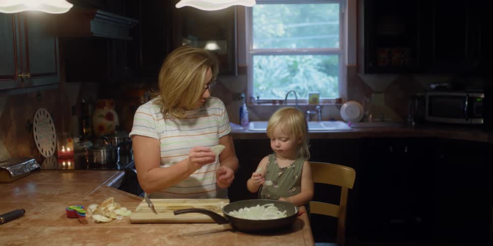 A young toddler stands on a chair and helps a woman with food in the kitchen