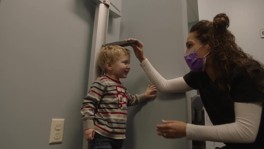 A boy and a nurse with a mask in a clinic with the nurse measuring the boys height next to a wall