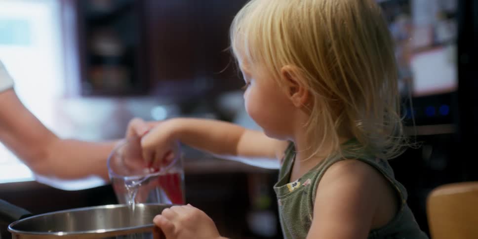 A woman pours water into a mixing bowl as a small toddler plays in the water pouring