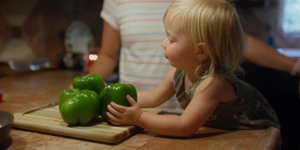 A little girl reaches for green peppers that are on a cutting board in a kitchen