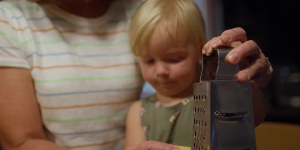 A woman grates a piece of cheese with a toddler watching
