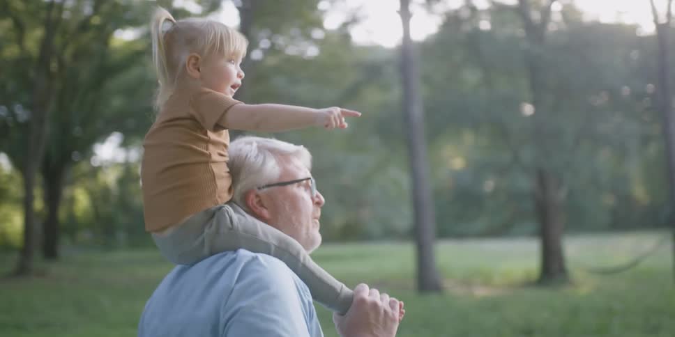 An older man holds a toddler girl on his shoulders as they walk through a rural grass area
