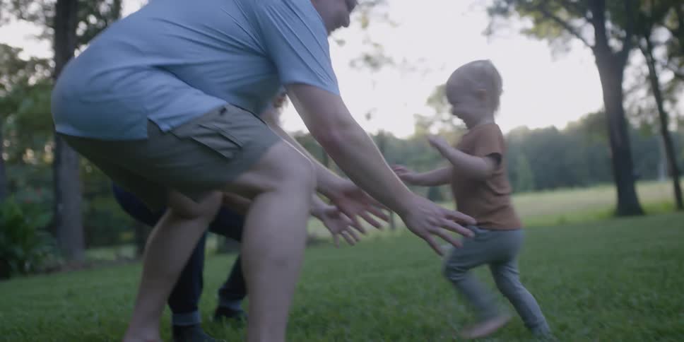 Blonde child runs across lawn to be picked up and hugged by grandparents with fields and woods in background
