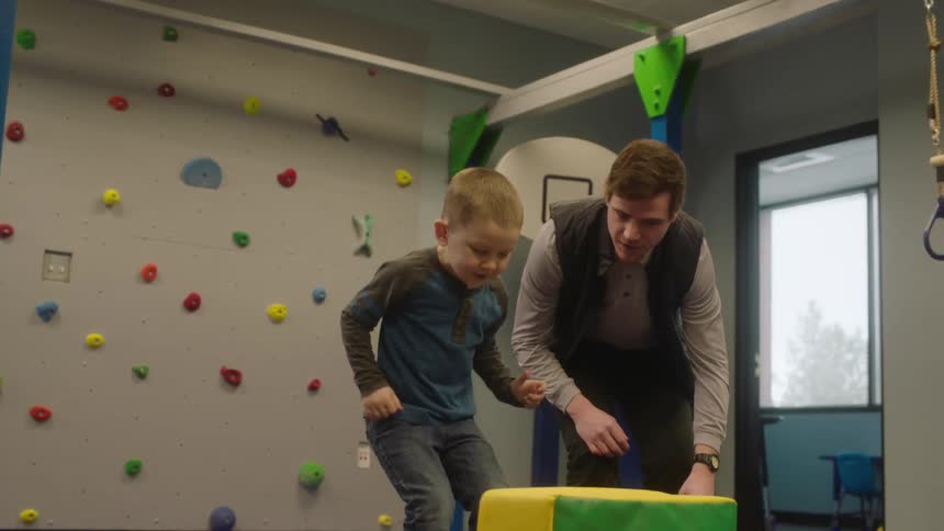A boy makes his way through an obstacle course in an inside gym while a man guides him
