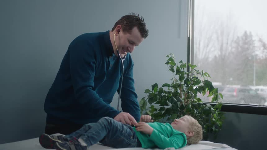 A man uses a stethoscope to listen to a toddler boys chest laying on a medical table