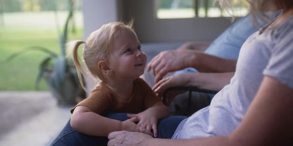 A toddler smiles as she hugs her moms knee as the family sits outside on their patio