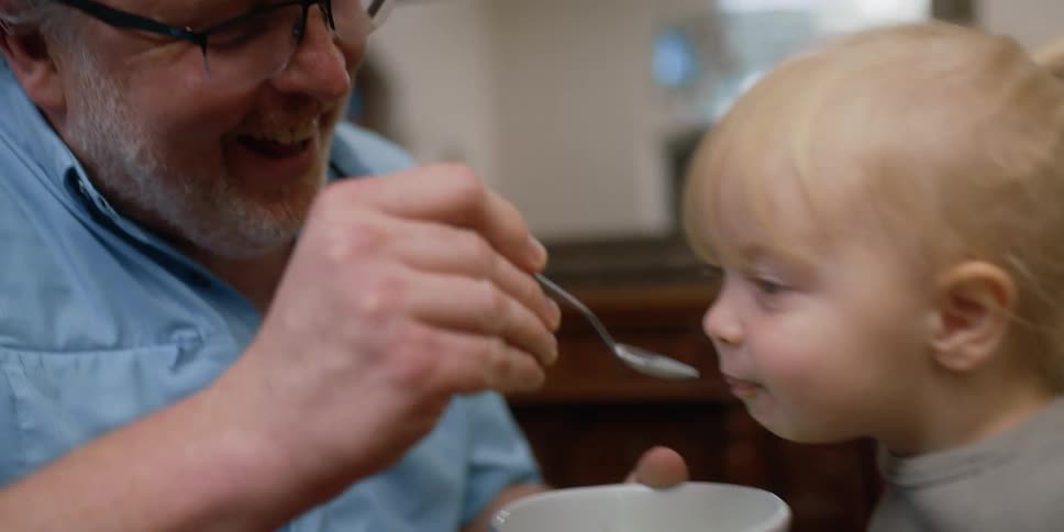 Grandfather with glasses and blue shortsleeved shirt feeds blonde granddaughter laughing