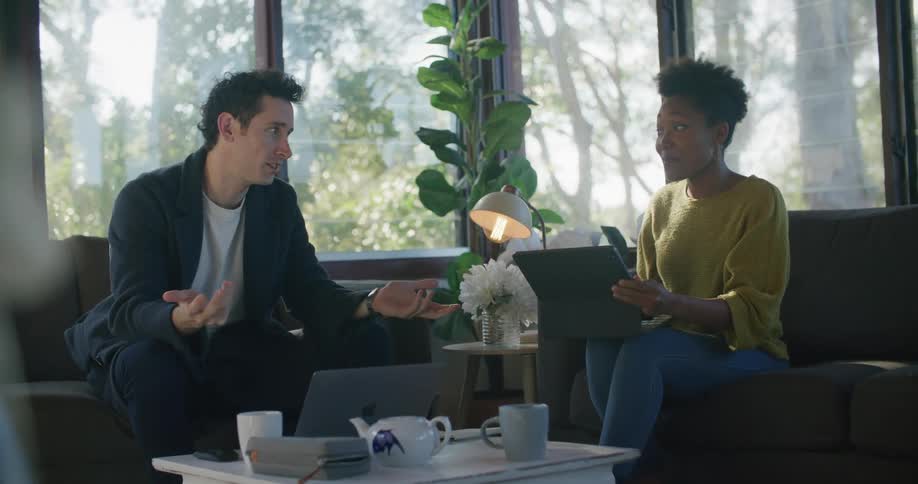 A man and a woman sitting on opposite sofas in a living room talk to one another while using their laptops
