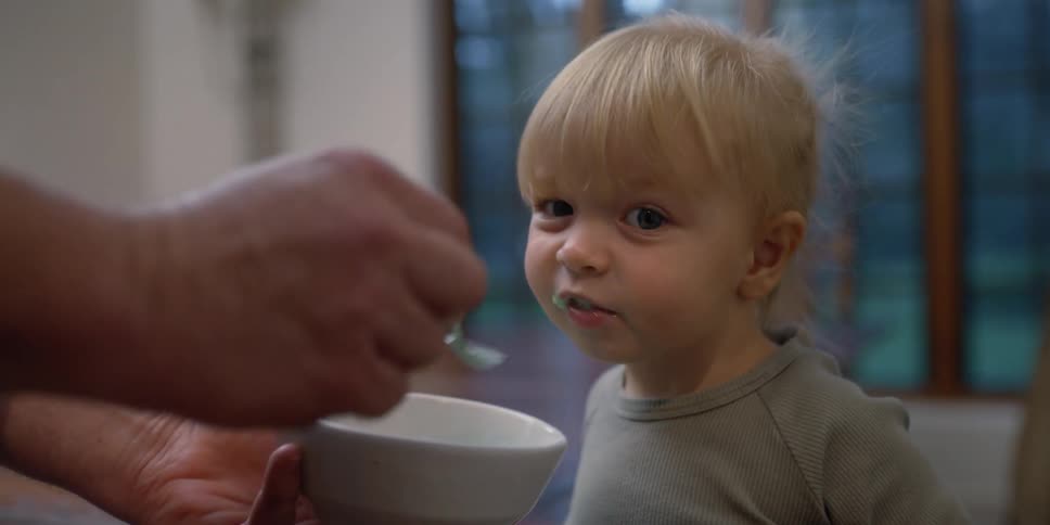 Person feeds blonde girl child from white bowl near dining table