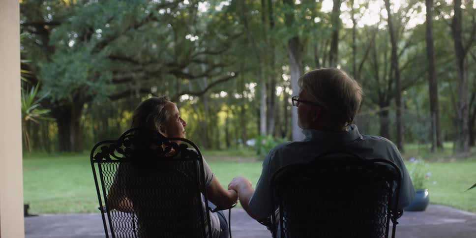 An elderly man and woman hold hands while sitting in rocking chairs in their yard during the day