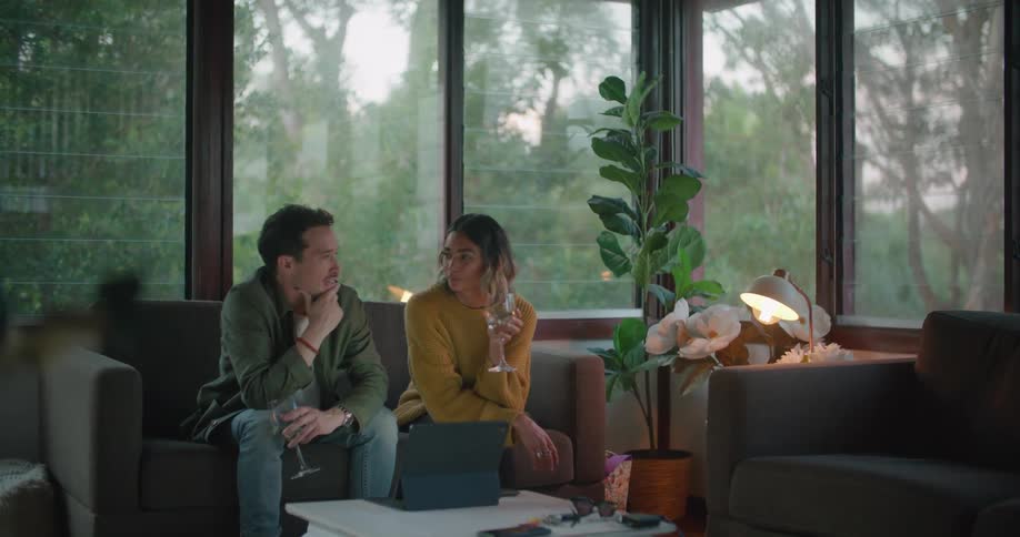 A man and a woman in a room sitting on a couch with a window and a plant in the background