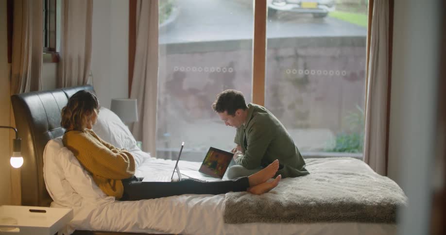 Woman reclines on a bed while a man sits near her and they talk while they both look at their laptops