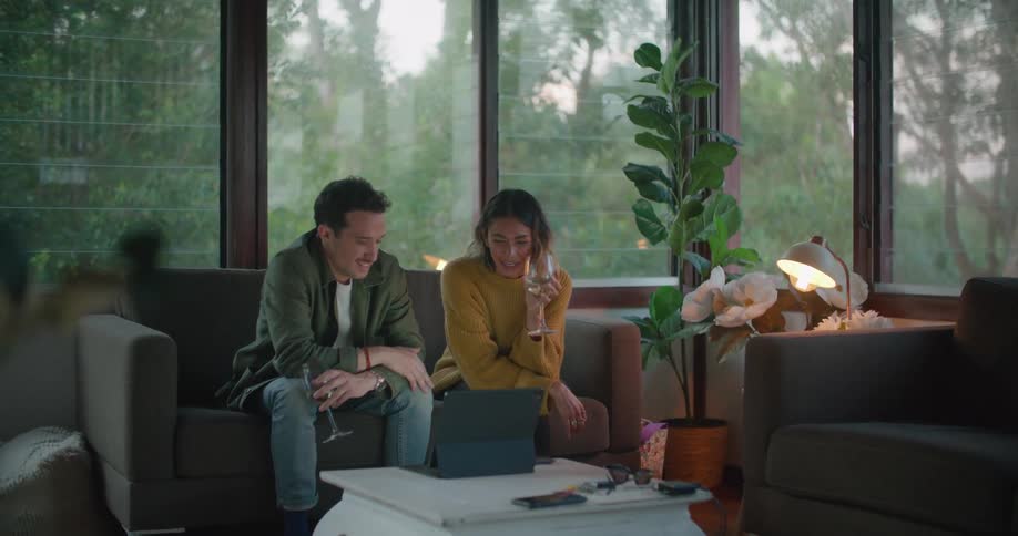 A man and woman chat and drink wine while watching the screen of a laptop in the living room of a house