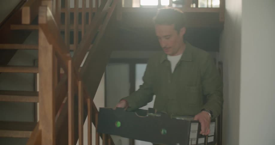 A man carrying a box and a woman carrying a bagged bottle of wine enter a house and ascend a wooden staircase