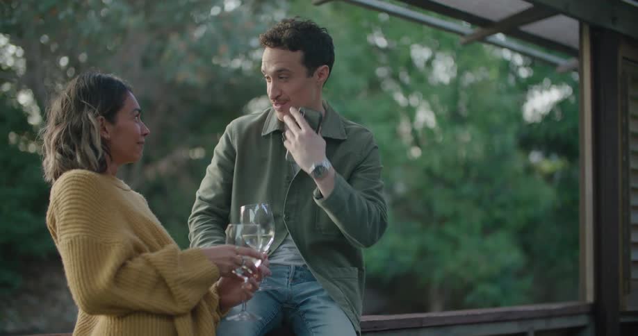 Man sits on porch rail holding wine glass and talking to woman