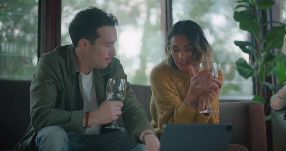 Couple sits on sofa drinking wine and talking near laptop