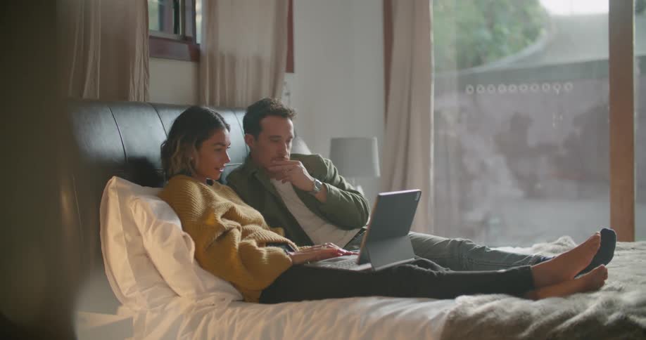 Couple reclines on bed sharing laptop with view of backyard