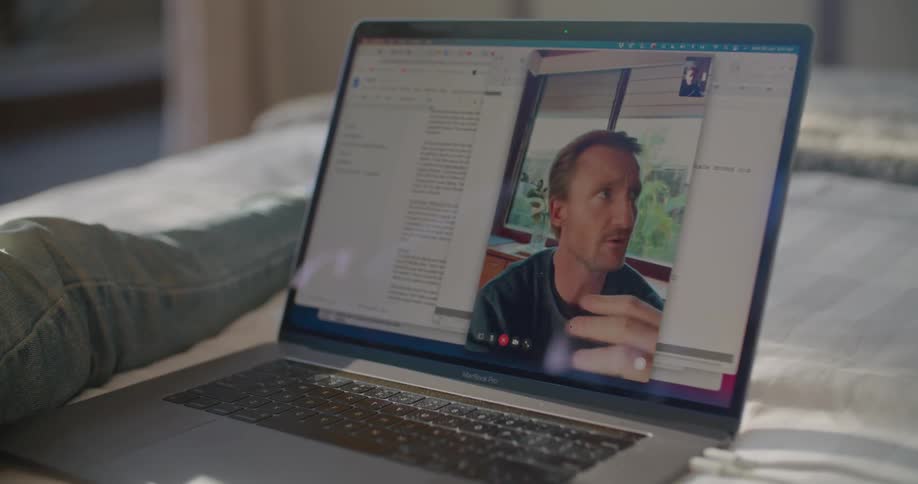 A man lying in bed talks to a friend via video call on his laptop