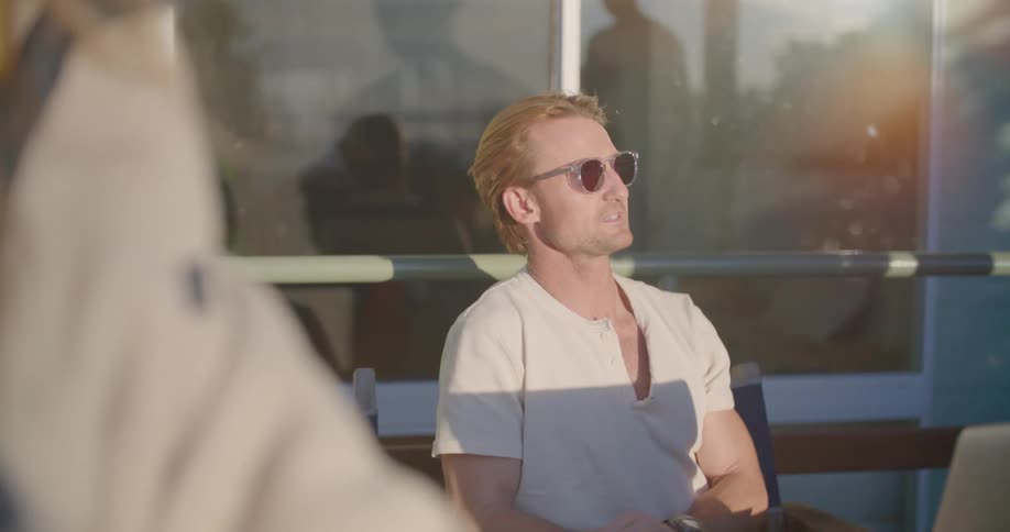 Man wearing shades and short sleeve shirt sits outdoors talking to man wearing suit reflected in building window