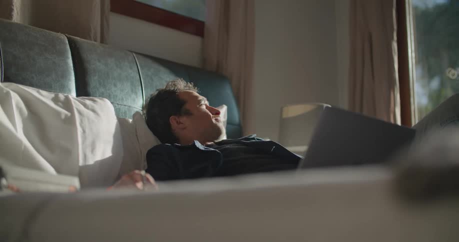 Man reclining on bed with laptop lies back looking outside and then up at the ceiling