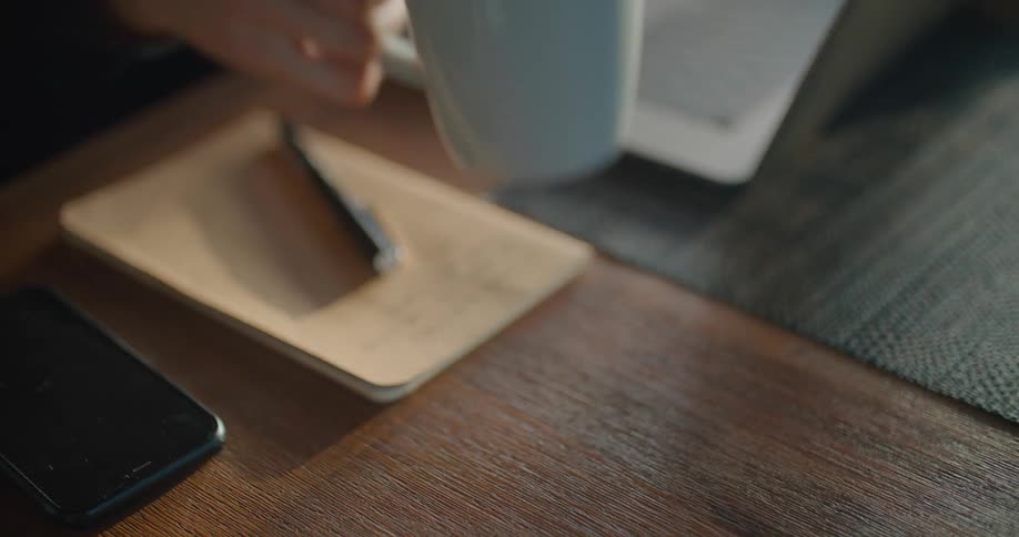A person sitting at a table with a smart phone and a laptop picking up a cup of coffee