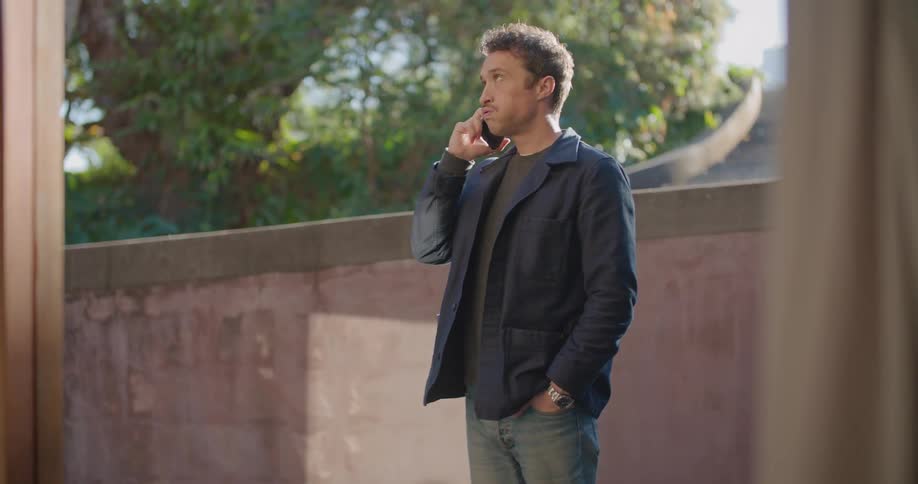 A man standing talking on a smartphone with a wall a driveway trees the sky and clouds in the background