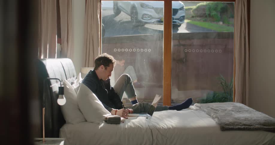 A man sitting on a bed writes in a notebook while using a laptop